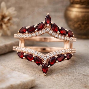 May include: A rose gold ring guard featuring two chevron-shaped bands. Each band is adorned with marquise-cut deep red garnet gemstones and small round white accent stones arranged in a pavé setting along the metal edges.
