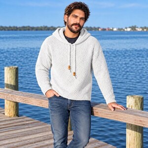 May include: A white long-sleeve hooded sweater with a textured knit pattern, featuring a dark gray crew neck undershirt, a hood with matching white drawstrings, and two small wooden beads attached to the ends of the drawstrings, paired with blue jeans.