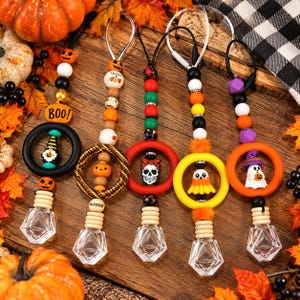 May include: Five hanging car air freshener diffusers featuring silicone and wooden beads in orange, black, white, and green. Each includes a circular ring, a decorative charm like a ghost, skull, or gnome, and a clear diamond-shaped glass bottle.