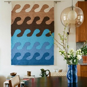 May include: A rectangular woven wall hanging featuring a repeating horizontal wave pattern in stacked bands of beige, dark brown, light blue, and dark blue, suspended from a silver metal rod mounted horizontally on a plain white wall.