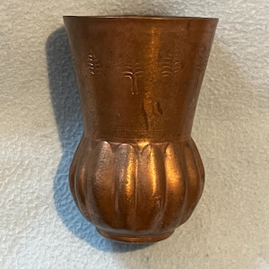 Puede incluir: Un jarrón de metal color cobre con un borde acampanado y una base redondeada y acanalada. La sección superior del jarrón presenta un patrón repetitivo en relieve de pequeños árboles estilizados que rodean la superficie exterior del recipiente.