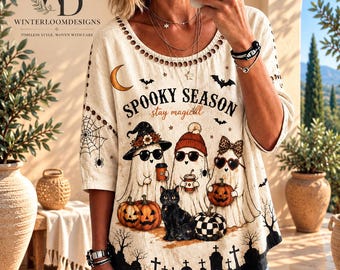 Puede incluir: Una blusa color crema con escote redondo y patrón recortado. El frente muestra el texto "SPOOKY SEASON" y "stay magical" sobre tres fantasmas con sombreros y gafas de sol, un gato negro, calabazas y la silueta de un cementerio.