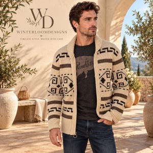 May include: A cream-colored zip-up cardigan featuring a shawl collar and a dark brown geometric pattern across the chest and sleeves. The knit garment is worn over a dark gray t-shirt with a graphic print and paired with dark blue denim jeans.