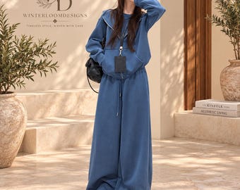Puede incluir: Un conjunto de descanso azul de dos piezas que consta de una sudadera con capucha, cremallera y mangas largas con bolsillo frontal, y pantalones anchos a juego con cordón. La sudadera incluye un cordón negro con tarjetero. Texto: WINTERLOOMDESIGNS.