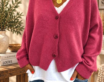 Puede incluir: Un cárdigan de color magenta de manga larga con un escote en V profundo y tres botones a juego en la parte delantera. El cárdigan se lleva sobre una camiseta blanca lisa y unos vaqueros azules. Un collar con colgante dorado descansa sobre el pecho.