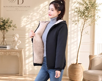 Peut inclure: Une veste à capuche noire avec une doublure en fausse peau de mouton beige, portée sur un pull à col roulé gris et un jean bleu. La veste présente une fermeture éclair frontale et des manches longues, avec le logo WinterloomDesigns en haut à gauche.