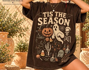 Puede incluir: Una camiseta de manga corta de color marrón oscuro con el texto 'TIS THE SEASON' en letras grandes de color crema. El gráfico incluye una calabaza, un fantasma, un esqueleto, un gato negro, un sombrero de bruja y pociones.
