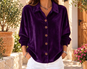 Puede incluir: Una camisa de manga larga de terciopelo color morado intenso con cuello clásico y cinco botones visibles en la parte delantera, combinada con pantalones blancos lisos. La camisa presenta una textura suave y afelpada con un corte holgado.