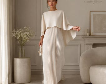 Puede incluir: Un vestido largo de color crema con falda plisada y una capa integrada que cae sobre los hombros. La prenda presenta un cuello redondo y una textura lisa y sólida en la parte de la capa, que contrasta con los pliegues verticales de la falda.