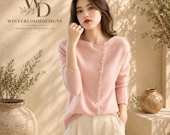 Puede incluir: Un suéter de manga larga en color rosa claro suave con una tapeta de botones diagonal con pequeños botones blancos, un escote ancho y un acabado de punto texturizado. El texto "WD WINTERLOOMDESIGNS TIMELESS STYLE, WOVEN WITH CARE" es visible.