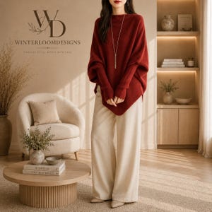 May include: A deep red long-sleeve sweater with a boat neckline and batwing sleeves, paired with cream-colored wide-leg trousers. The sweater features a long gold-tone pendant necklace. The logo WD WINTERLOOMDESIGNS is visible in the upper left corner.