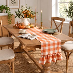 May include: An orange and white gingham table runner with a ruffled edge, draped over a wooden dining table. The runner features a classic checkered pattern and is styled with a white ceramic pitcher, small pumpkins, and two tall taper candles.