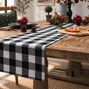 May include: A rectangular table runner featuring a classic black and white buffalo check pattern. The fabric has a woven texture and is laid flat across the center of a wooden dining table, extending from the edge toward the center of the table.
