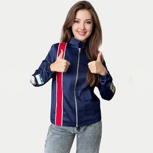 Könnte beinhalten: Eine marineblaue Reißverschlussjacke mit einem vertikalen roten Streifen auf der rechten Seite, einem Stehkragen und kleinen rechteckigen Aufnähern an beiden Ärmeln. Die Jacke hat einen silberfarbenen Frontreißverschluss und eine kleine Tasche.