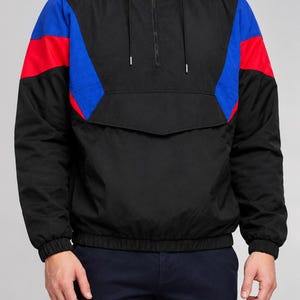 Puede incluir: Una chaqueta cortavientos negra tipo pullover con cierre de media cremallera, capucha con cordón y mangas largas. El diseño incluye paneles de bloques de color azul y rojo vibrantes en el pecho y los hombros, con puños y dobladillo elásticos.