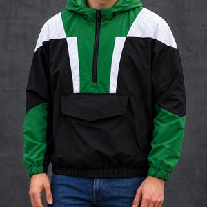 May include: A colour-blocked pullover hoodie featuring a green hood and upper chest, black lower body and sleeves, and white accents on the shoulders and chest. The garment includes a half-zip front closure and a large horizontal front pocket.