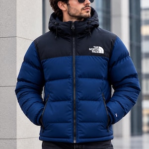 May include: A blue and black puffer jacket with a hood, featuring a full-length front zip, two zipped side pockets, and the white text logo THE NORTH FACE printed on the upper left chest area of the black yoke section of the garment.