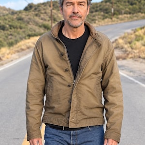 May include: A tan canvas jacket with a brown corduroy collar, a front zip, and button fastenings, worn over a black t-shirt with blue denim jeans. A green stone ring is visible on one finger, and the garment features long sleeves and pockets.