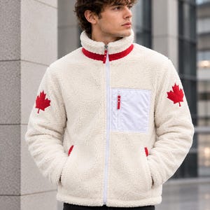 Op de afbeelding: Een crèmekleurig sherpa fleecejack met een hoge kraag met rode bies, een witte ritssluiting over de hele lengte en een witte borstzak. Op beide mouwen zijn rode esdoornblad-patches geborduurd en de zakken zijn voorzien van rode accenten.