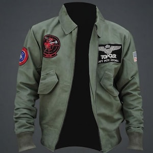 Puede incluir: Una chaqueta tipo bomber de color verde oliva con cuello negro, puños acanalados y cremallera frontal. Los parches incluyen un emblema circular rojo, un parche rectangular negro con el texto TOP GUN CAPT PETE MITCHELL y una pequeña bandera de EE. UU.