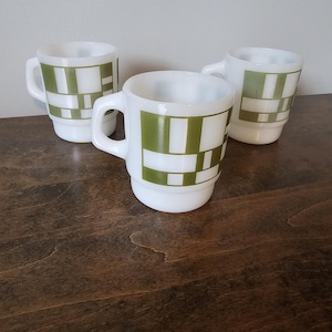 Op de afbeelding: Drie mokken van wit melkglas met een geometrisch patroon van elkaar kruisende olijfgroene rechthoeken en vierkanten. Elke mok heeft een afgerond handvat en een stapelbaar ontwerp, met een consistent herhalend rastermotief aan de buitenkant.