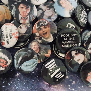 May include: A collection of circular pin-back buttons featuring various graphic designs, including portraits of a person, text such as 'POOL BOY AT THE VAMPIRE MANSION' and 'DEAD!', and band-related imagery, all arranged on a dark, starry background.