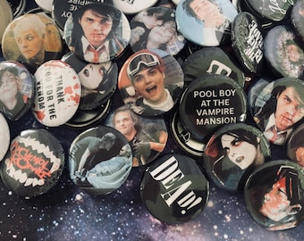 May include: A collection of circular pin-back buttons featuring various graphic designs, including portraits of a person, text such as 'POOL BOY AT THE VAMPIRE MANSION' and 'DEAD!', and band-related imagery, all arranged on a dark, starry background.