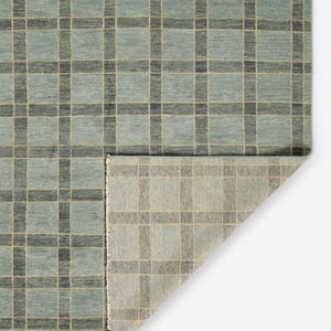 May include: A rectangular area rug featuring a grid pattern of dark charcoal lines over a muted sage green background. The rug is folded at one corner, revealing a lighter, beige-toned underside with a visible woven texture and fringe edge.