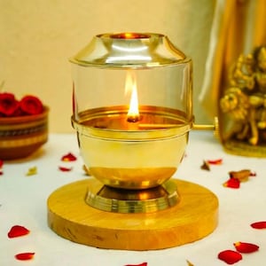 Può includere: Una lampada a olio in ottone con un cilindro in vetro trasparente e una fiamma accesa all'interno. La lampada presenta una finitura color oro metallizzato, una base circolare e una piccola manopola di regolazione laterale.