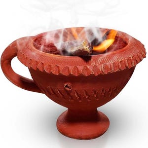 May include: A terracotta-colored clay incense burner with a pedestal base and a side handle. The bowl features a textured, notched rim and carved geometric patterns on the exterior. A piece of burning wood emits smoke and a small flame from the center.