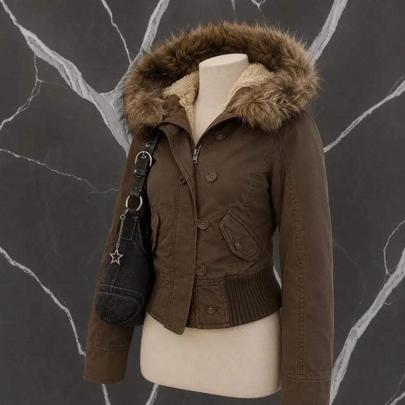 May include: A brown jacket with a hood featuring a brown faux fur trim and a beige fleece lining. The jacket has a button-up front, long sleeves, and a ribbed hem. A black shoulder bag with a silver star charm is hanging from the jacket strap.