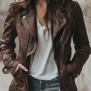 May include: A dark brown leather jacket with a wide lapel, silver-toned metal zippers, and snap button details on the cuffs and collar. The jacket is worn over a plain white t-shirt and distressed blue denim jeans with visible rips.
