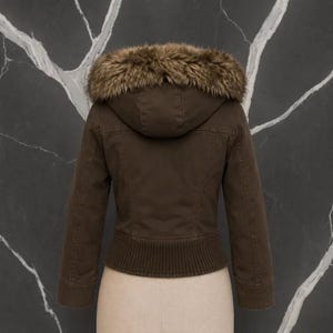 May include: A dark brown jacket featuring a hood trimmed with thick, textured faux fur in shades of brown and tan. The jacket has long sleeves, a ribbed knit hemline, and visible seam detailing across the back, providing a structured fit.