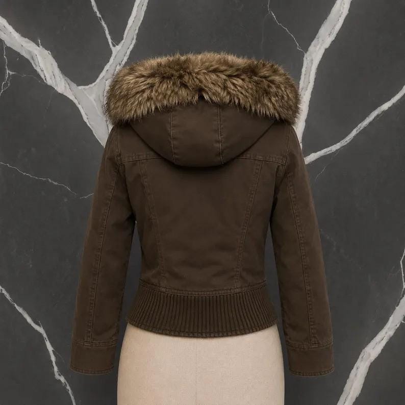May include: A dark brown jacket featuring a hood trimmed with thick, textured faux fur in shades of brown and tan. The jacket has long sleeves, a ribbed knit hemline, and visible seam detailing across the back, providing a structured fit.