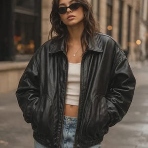 May include: A black leather bomber jacket with a collared neckline, long sleeves, and a front zipper closure, worn over a white cropped tank top and light blue denim jeans, paired with dark oval-shaped sunglasses and a silver chain necklace.