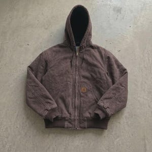 May include: A brown hooded zip-up jacket featuring a black interior lining, a front zipper closure, two lower front pockets, and a small rectangular tan brand patch on the right pocket. The jacket has ribbed cuffs and a ribbed hem at the waist.