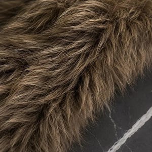May include: A dense, long-pile faux fur fabric in shades of brown and tan with a shaggy, multi-tonal texture. The fibers are layered and oriented in various directions, creating a realistic, voluminous appearance with light and dark highlights.