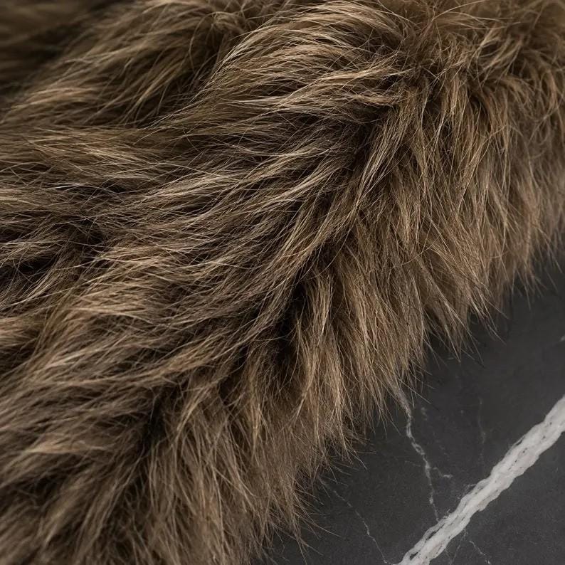 May include: A dense, long-pile faux fur fabric in shades of brown and tan with a shaggy, multi-tonal texture. The fibers are layered and oriented in various directions, creating a realistic, voluminous appearance with light and dark highlights.
