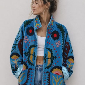 Puede incluir: Una chaqueta de terciopelo azul brillante con intrincados bordados florales multicolores en tonos rojo, amarillo y verde oscuro. La chaqueta tiene cuello alto, mangas largas con puños a rayas azul claro y blanco, y cierre de cremallera frontal.