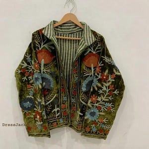 May include: An olive green velvet jacket featuring intricate floral embroidery in shades of orange, blue, and white. The jacket has a collared neckline and an interior lining with a green and white vertical striped pattern. The text DressJack is visible.