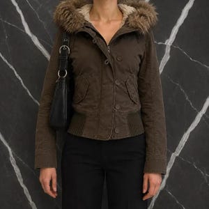 May include: A dark brown jacket featuring a hood with a faux fur trim, a front button closure, and ribbed knit detailing at the hem. The jacket includes two front pockets with flaps and long sleeves. A black shoulder bag is worn on the right side.