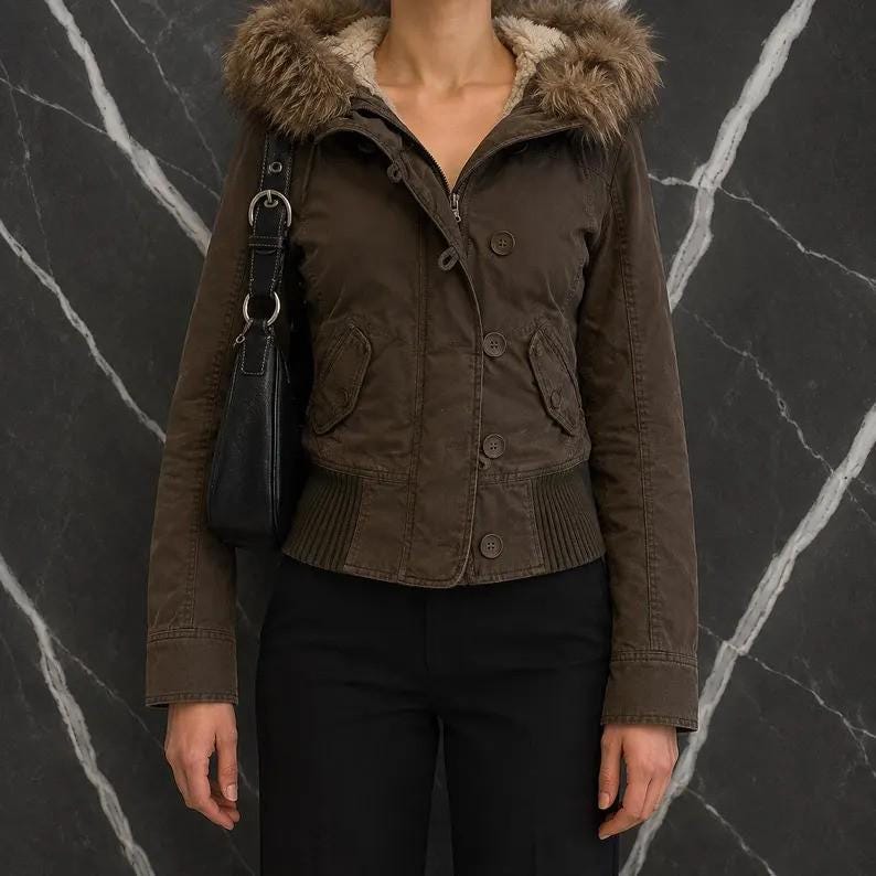 May include: A dark brown jacket featuring a hood with a faux fur trim, a front button closure, and ribbed knit detailing at the hem. The jacket includes two front pockets with flaps and long sleeves. A black shoulder bag is worn on the right side.