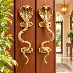 May include: Two brass-coloured metal door handles shaped like upright cobras with flared hoods and detailed scale textures. Each handle is mounted to a dark wood door with two oval plates secured by visible screws, featuring an intricate pattern.