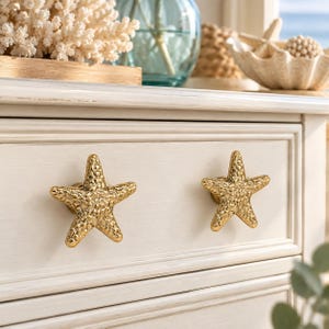 May include: Two gold-toned metal drawer knobs shaped like five-pointed starfish with a textured, bumpy surface. The knobs are attached to a cream-colored wooden drawer front with horizontal paneling and a distressed finish.