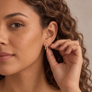 May include: A pair of gold-toned stud earrings featuring two thin, parallel vertical bars. The earrings are worn in the earlobe, showcasing a simple, clean design with a polished metallic finish that catches the light.