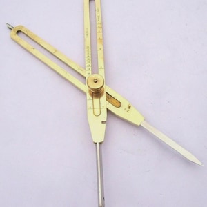 May include: A brass drafting compass with two intersecting arms featuring a central adjustment knob. The tool includes engraved markings labeled CIRCLES and FINE LINES, with numerical scales ranging from 1 to 20, and sharp metal points at the ends.