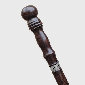 May include: A dark brown wooden walking stick featuring a smooth, rounded spherical handle top. The shaft includes a series of horizontal ridges below the handle and a decorative silver-colored metal band with an intricate woven pattern.