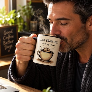 Könnte beinhalten: Ein weißer Keramikbecher mit schwarzem Rand und Henkel, verziert mit einer Grafik eines Gehirns in einer Kaffeetasse. Der Text lautet: LAST BRAIN CELL, Currently being caffeinated. Der Becher wird in der Hand gehalten, Dampf steigt daraus auf.