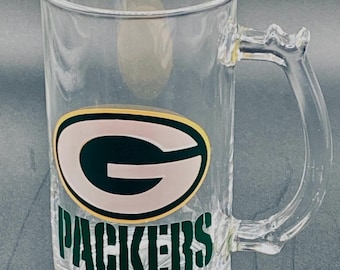 May include: Clear glass beer mug with a handle. The mug features the Green Bay Packers logo in green, gold, and white, with the word "PACKERS" in green below the logo. The mug has a textured base.