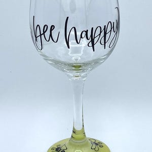 May include: A clear wine glass with the words "bee happy" in black script. The base of the glass is painted yellow with black bee and flower designs. The glass is designed for beverages.